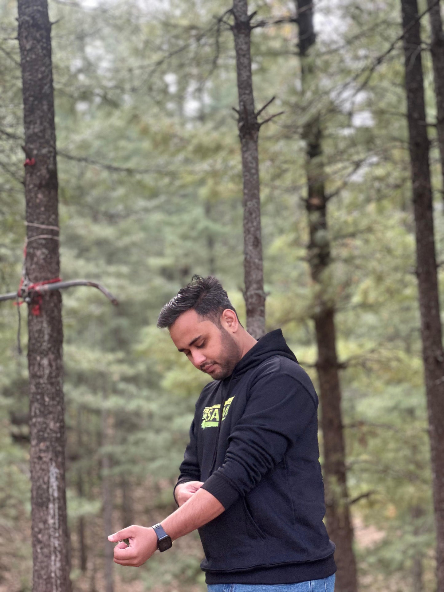 Black hoodie with 'SARIM' text design, worn by a model adjusting sleeves in a forest. Comfortable and stylish hoodie for everyday wear.