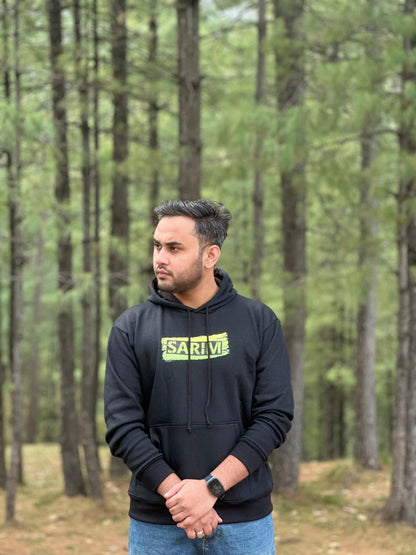 Black hoodie with 'SARIM' text design on the front, worn by a model in a forest. High-quality fabric and stylish fit for casual wear.