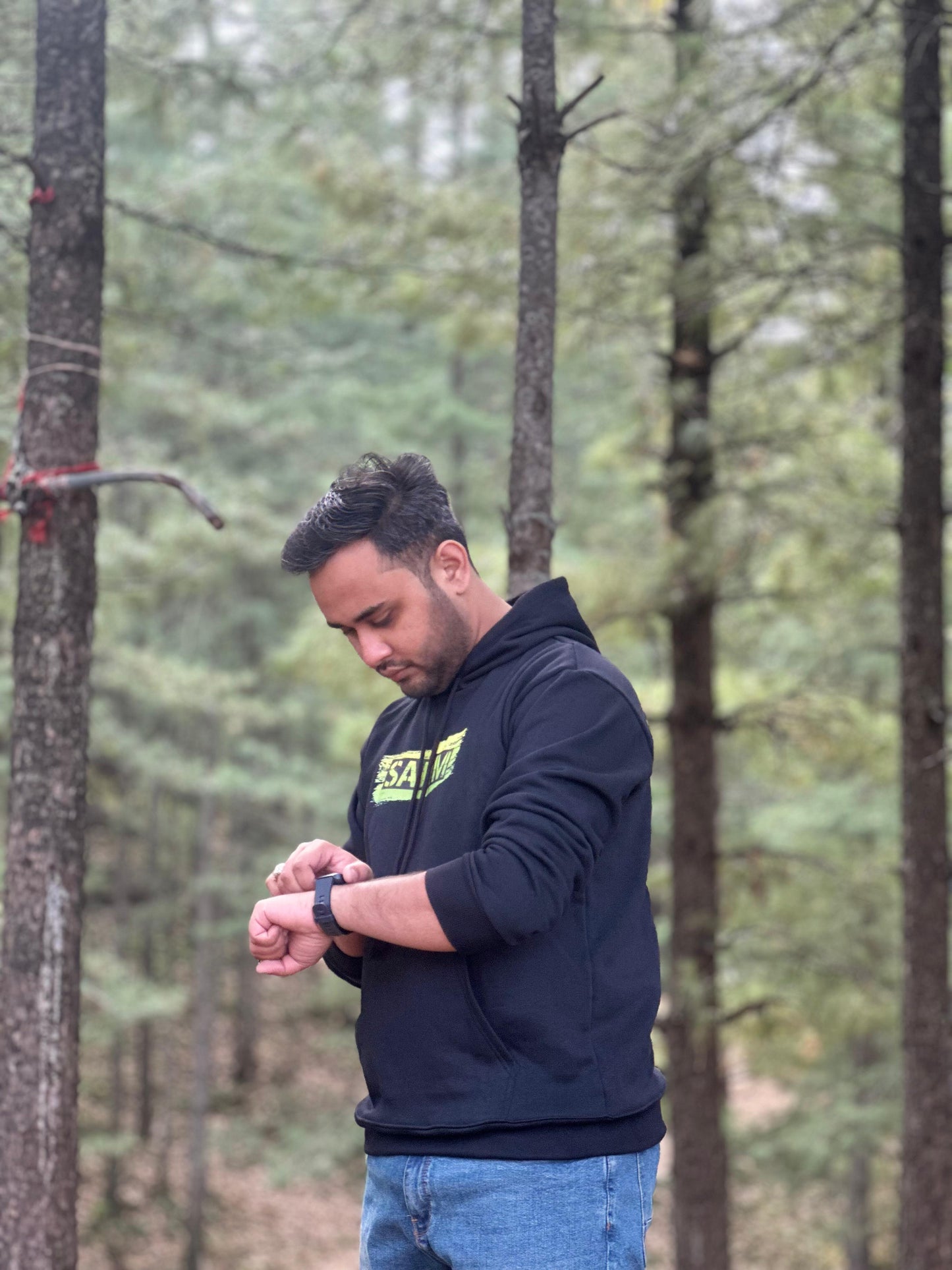 Black hoodie with 'SARIM' text design, worn by a model checking the time on a smartwatch in a forest. Trendy and cozy streetwear fashion.