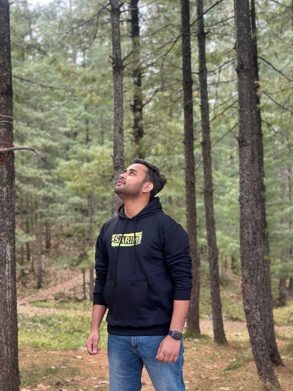 Black graphic hoodie with 'START' text design on the front, worn by a model in a natural outdoor setting. Trendy and cozy streetwear fashion.